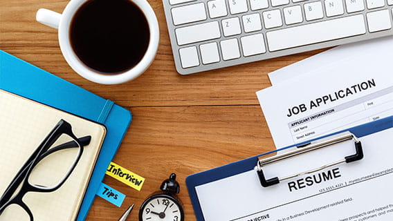 desk with a job application, resume, keyboard, coffee, glasses, pen, and clock 