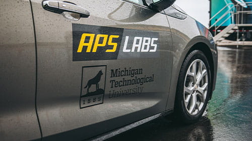 Close-up of a gray vehicle door displaying APS Labs and Michigan Technological University decals.