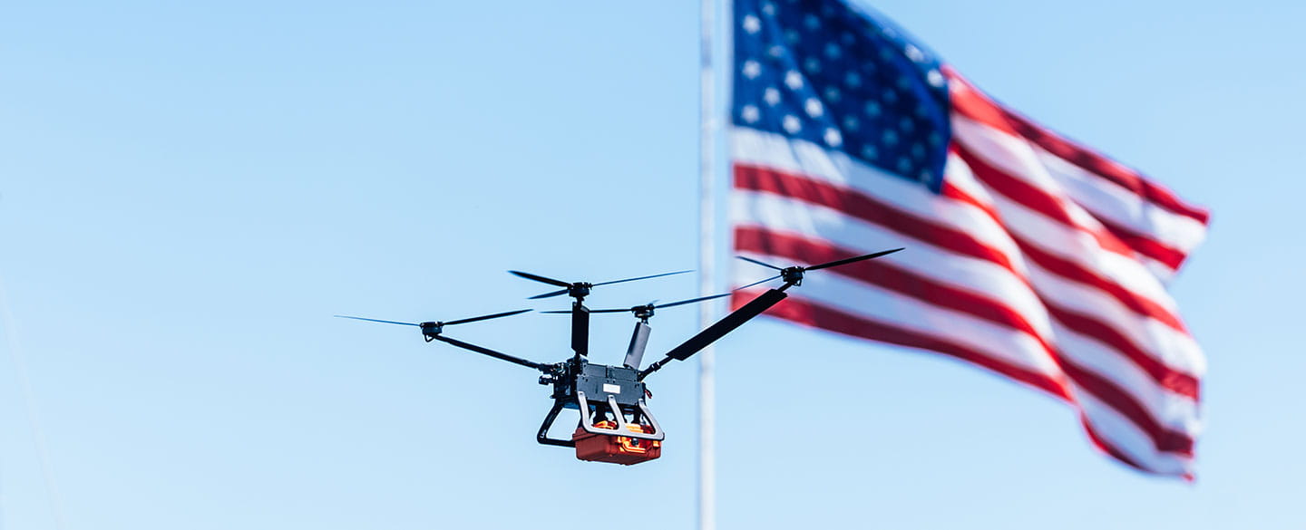 Drone carrying a package in front of an American flag.