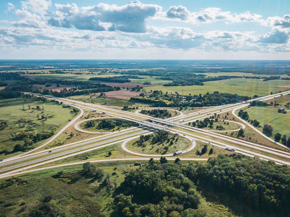 Aerial view of a highway interchange surrounded by rural fields and trees under a cloudy sky.