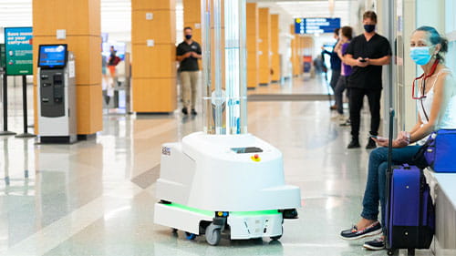Autonomous service robot moving through an airport terminal as travelers wait nearby.