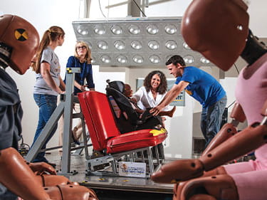 Researchers conducting automotive safety testing around a vehicle seat surrounded by crash test dummies.