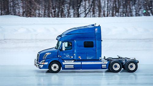 Blue semi-truck cab driving on an icy test track surrounded by snowbanks.