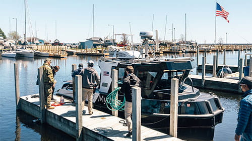 People standing on a marina dock beside a research boat with sailboats and an American flag in the background.