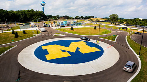 Aerial view of Mcity test facility with vehicles driving around a circular roadway featuring a large University of Michigan logo.