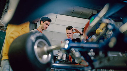 Two people working on a vehicle component in a workshop, viewed through mechanical parts in the foreground.