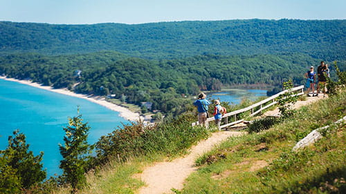 People walking along a scenic trail overlooking a blue lake and forested shoreline.