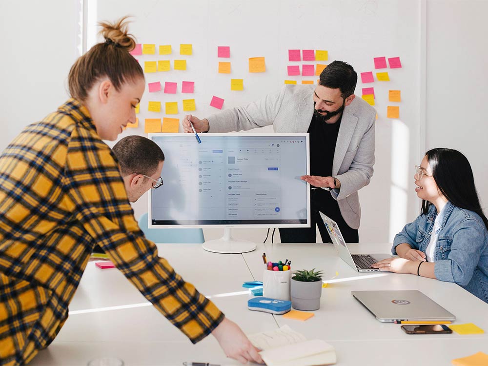 Team members collaborate around a computer monitor during a planning meeting with sticky notes on the wall.