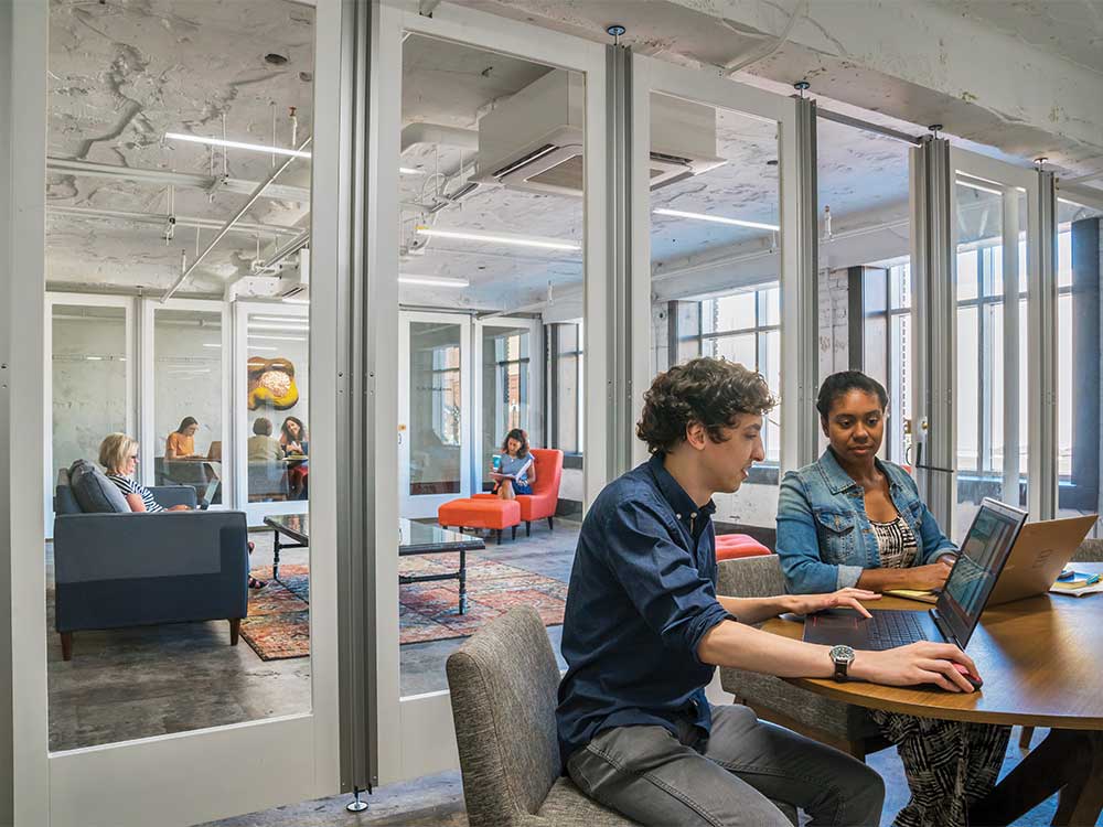 Two people work on laptops at a table in a modern glass-walled coworking space.