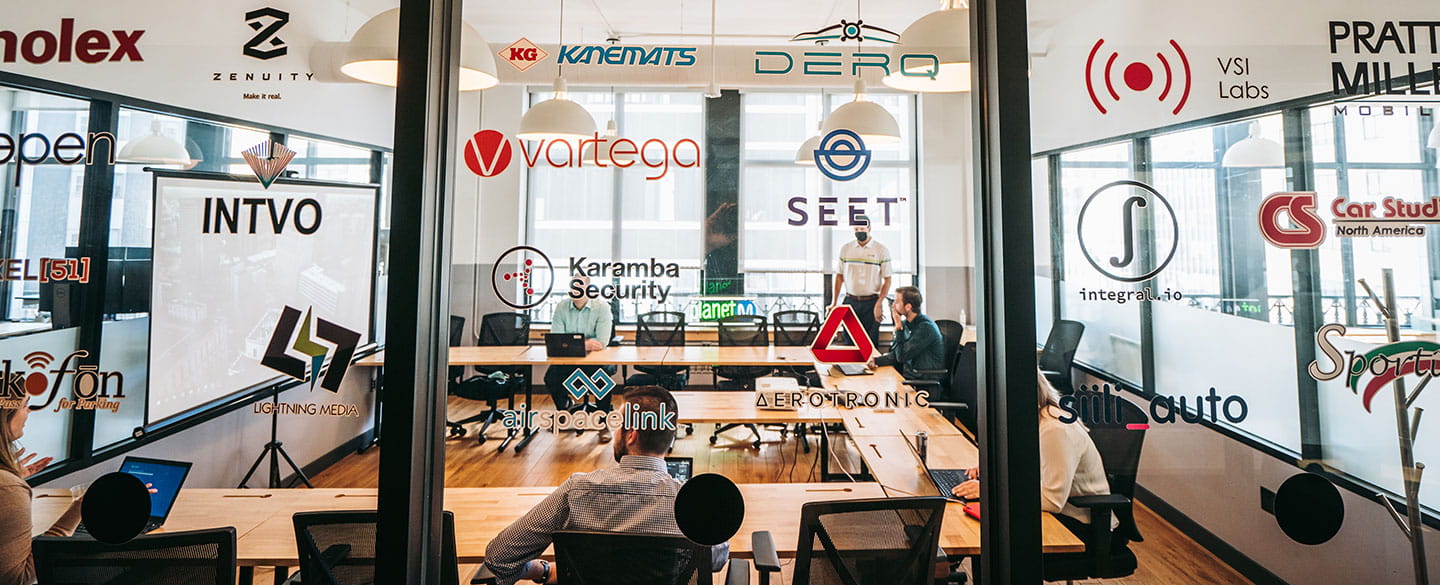 Company logos displayed on glass walls overlooking people working in a shared office and meeting space.