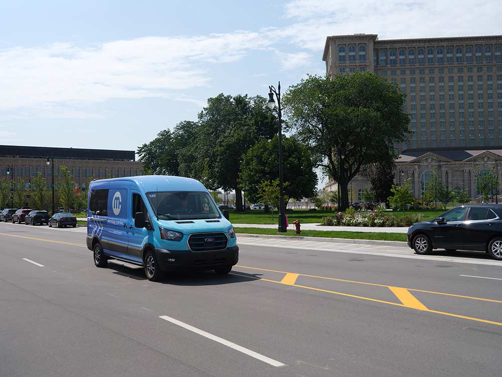 A blue Michigan Central electric van drives over wireless charging equipment embedded in a Detroit roadway.