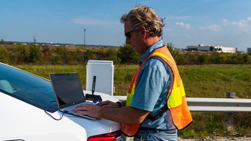 A worker in a safety vest uses a laptop beside a vehicle on a road shoulder.