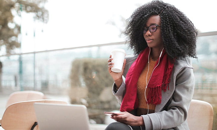 Black woman sitting in front of a laptop holding phone in one had and coffee cup in the other