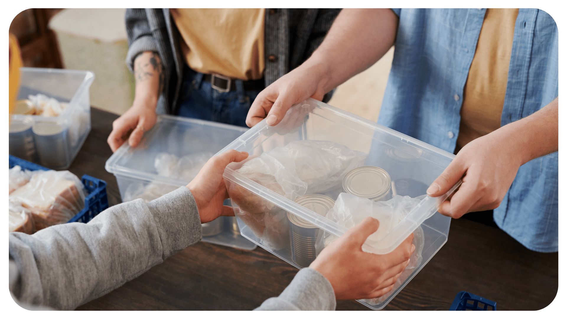 A pair of hands is handing another pair of hands a plastic tub with canned goods in it.