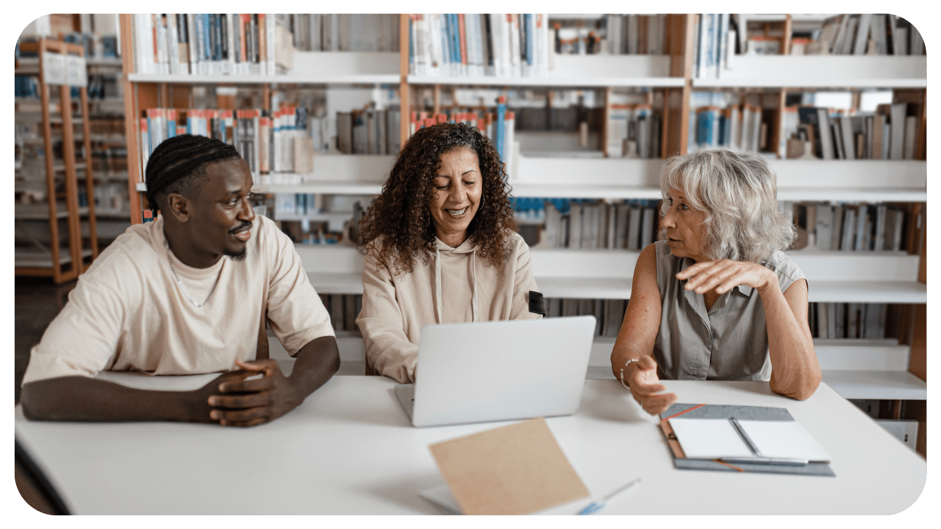 Three people sit in a library and are looking at each other smiling. The person in the middle is using a silver laptop.