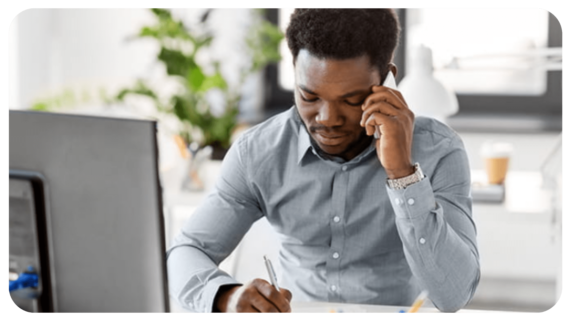 A person is on the phone and is looking down while writing something. There is a computer in front of them.