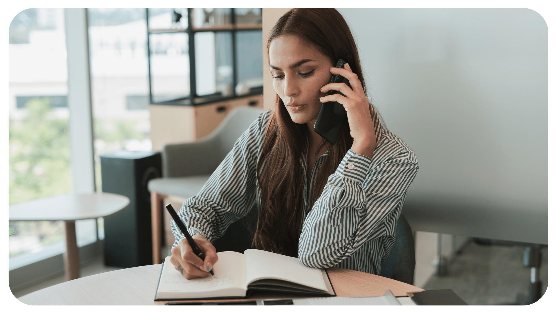 A person sits holding a smartphone to their ear while also writing at a desk.