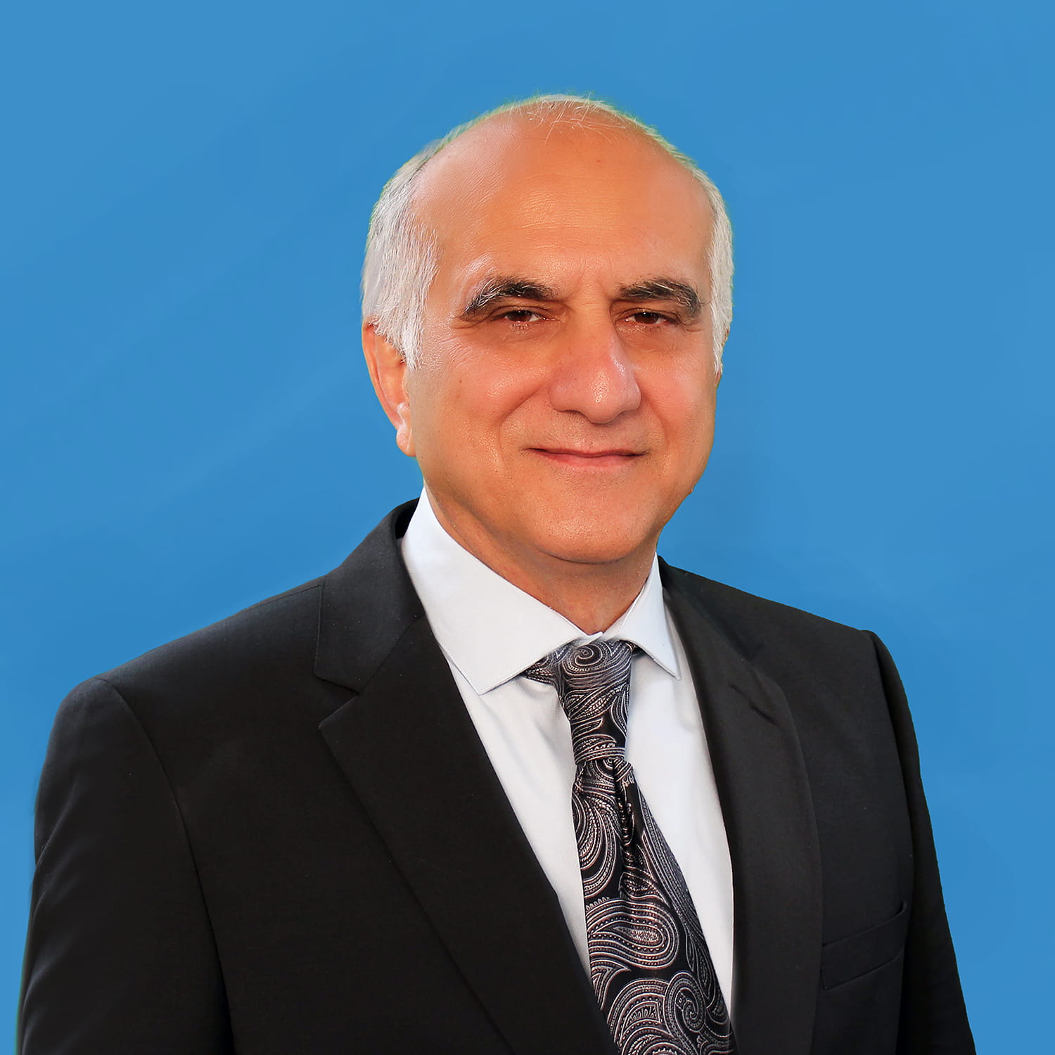 Head and shoulders photo of a balding, older white man, who is smiling into the camera. He is wearing a light blue shirt, dark tie, and a blazer. He has white hair on the sides of his head, the top of which is bald.