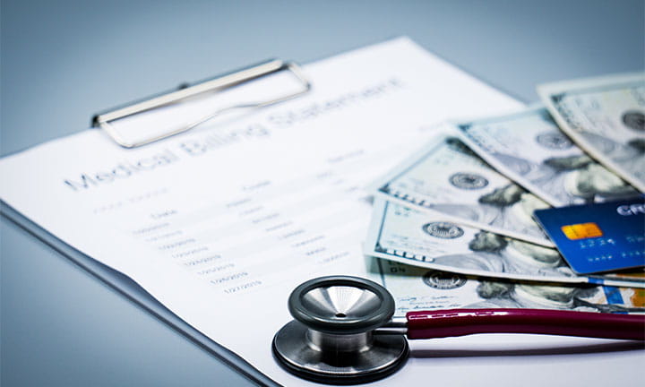 A clipboard holding a medical bill with money, a credit card, and a stethoscope on top of it