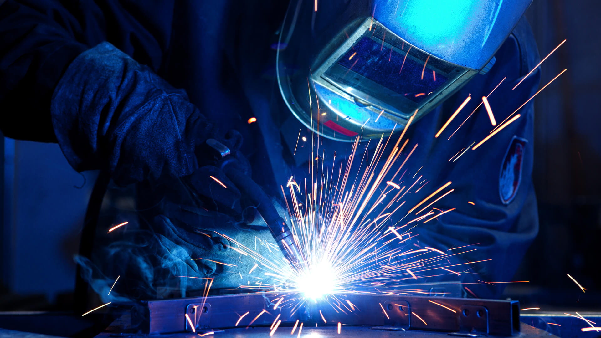 A metal worker wearing a welding mask and using a welding torch to fuse metal pieces while sparks project outward.