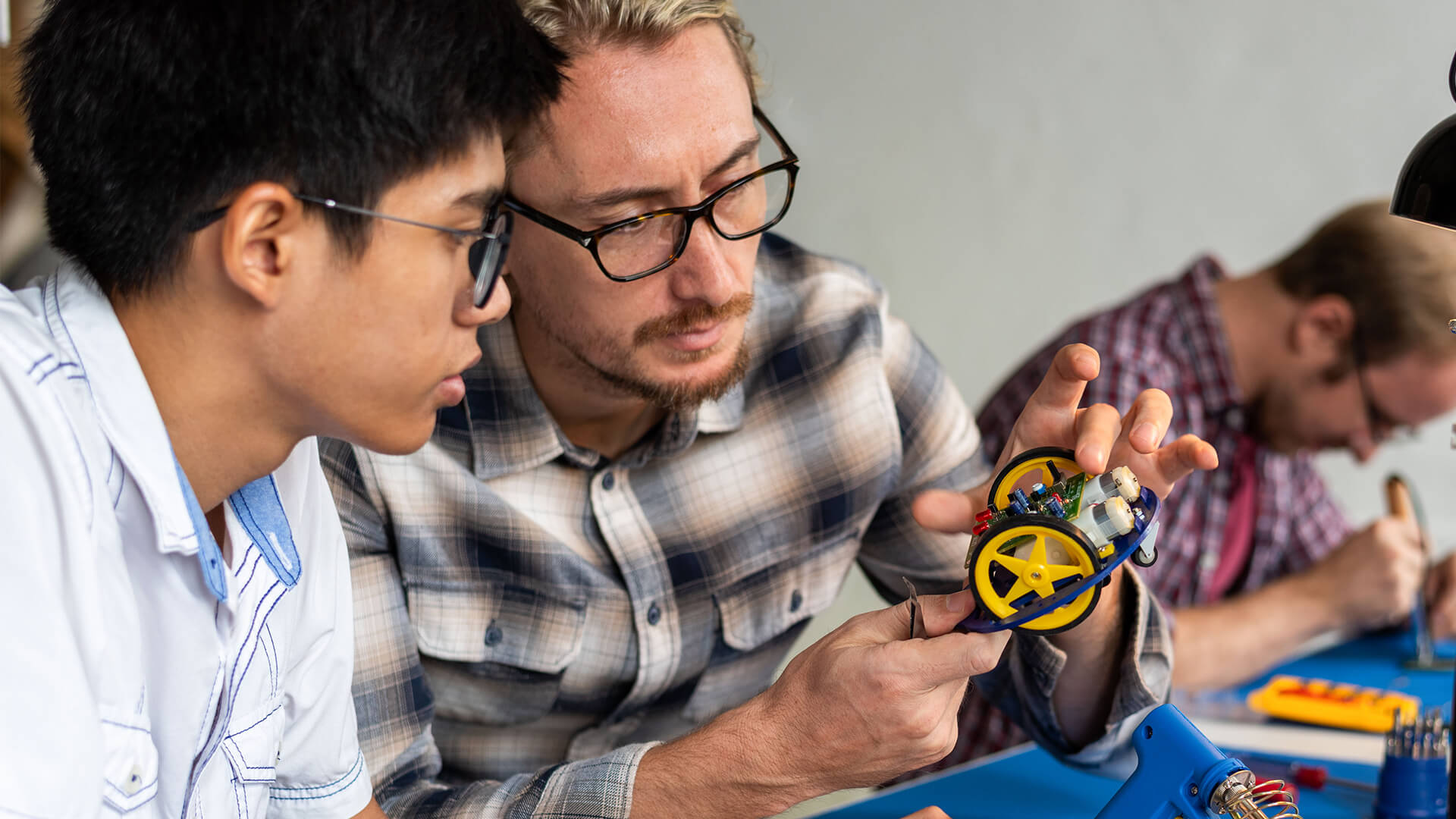 Close-up of a teacher guiding a student as they examine and assemble a small wheeled robot, pointing out electronic components while another student works in the background at a desk with tools.