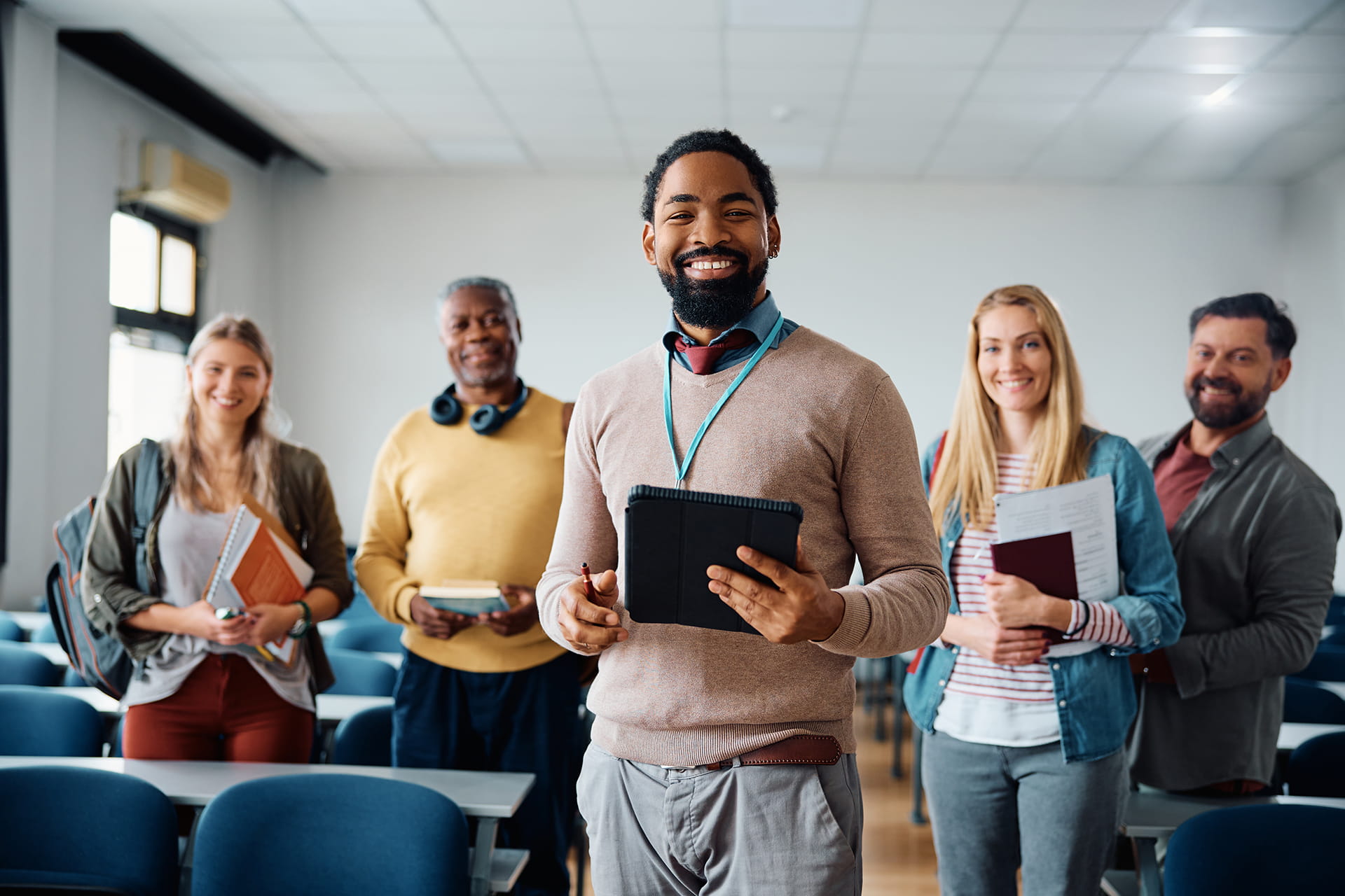 Adult learners standing together in a classroom, holding books, notebooks and a tablet.