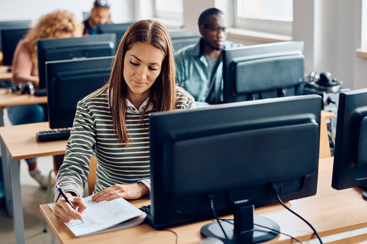 Middle-aged person sitting at a computer and writing in a notebook on a tabletop while other people use computers in the background.