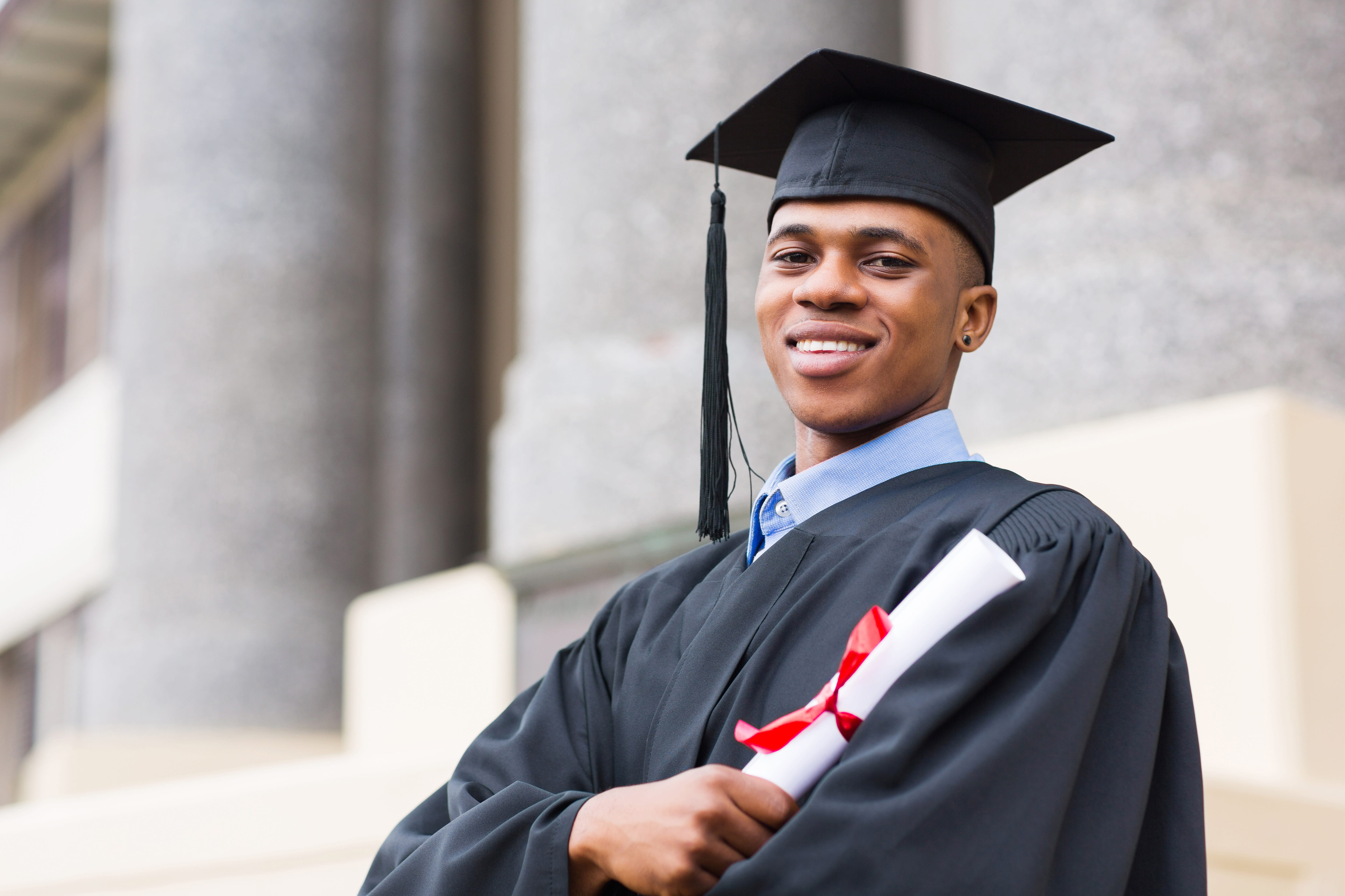 Man in a black cap and gown holding a rolled diploma in his crossed arms.