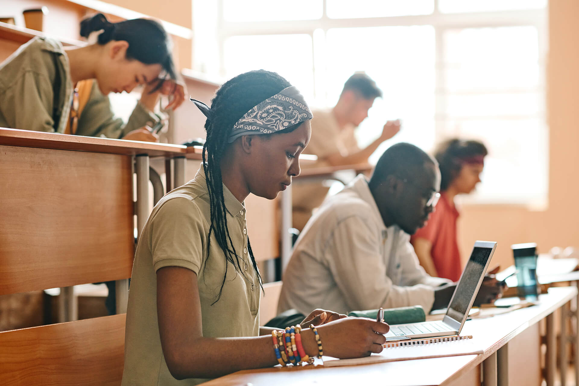 College students seated in a brightly lit stadium-style lecture hall working individually on their phones, laptops, and in notebooks.