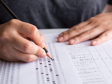 Student's hand writing with a pencil to fill in a bubble answer sheet next to a separate paper showing mathematical equations.