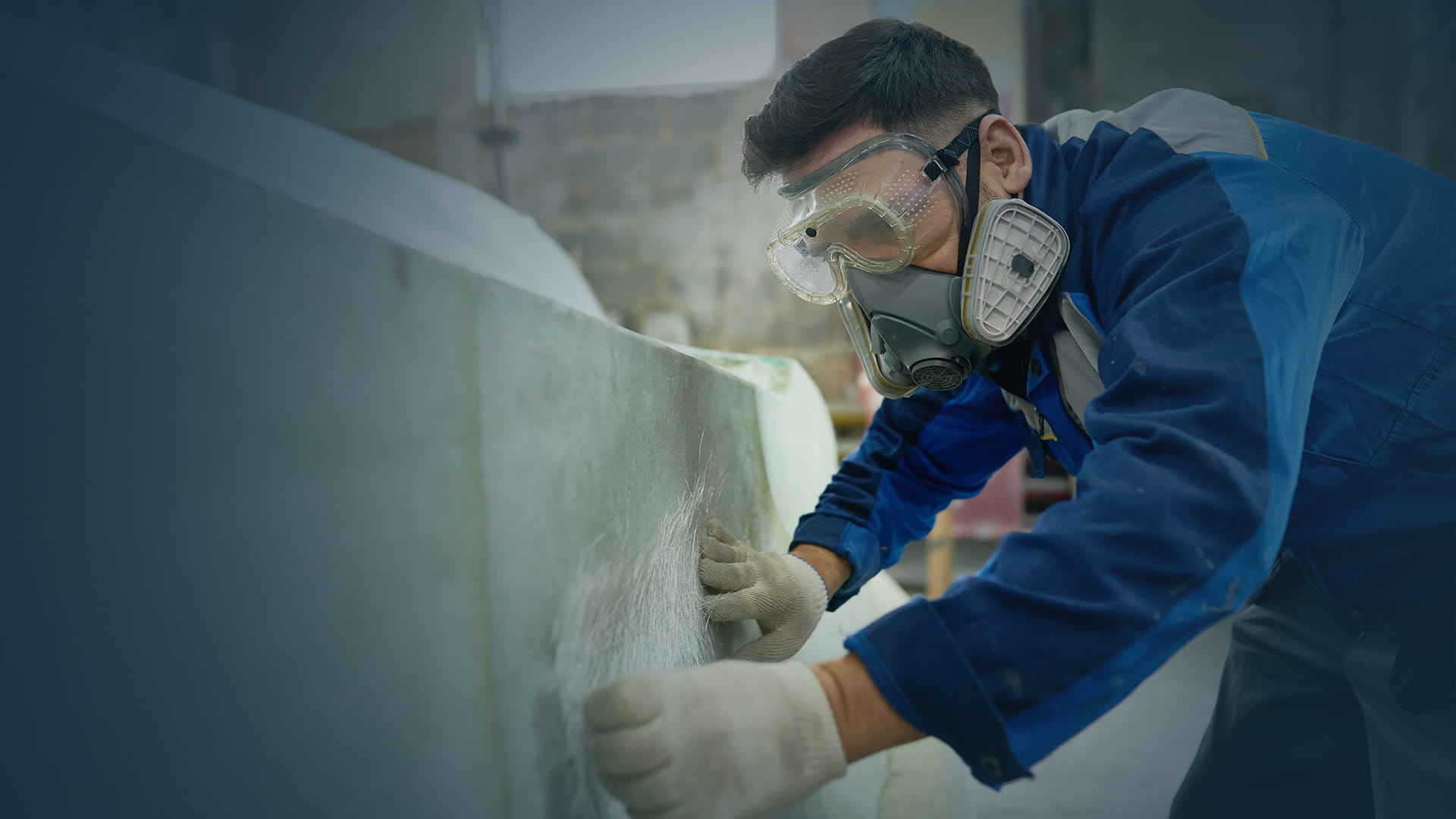 Worker wearing a respirator, safety goggles, and gloves applies a piece of fiberglass mesh onto a curved surface in a workshop.