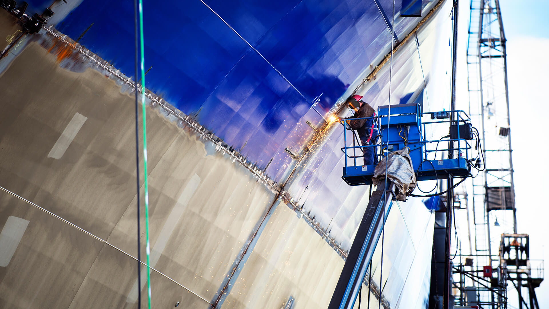 Worker in a lift welding a large blue metal ship hull, with bright sparks visible and industrial structures in the background.