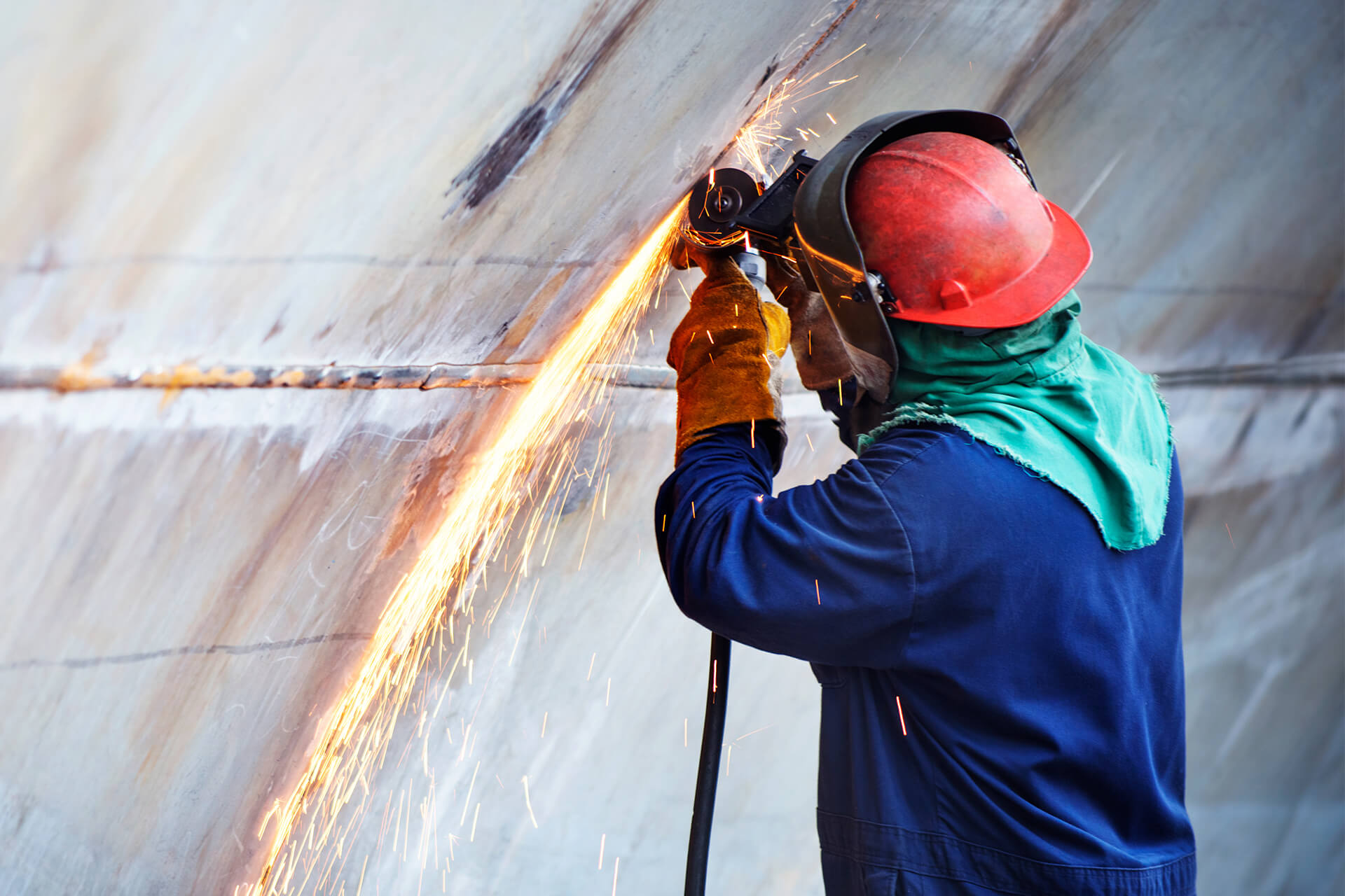 Worker wearing a red hard hat, face shield, gloves, and protective clothing grinds a weld seam on a large curved steel surface, sending sparks downward.