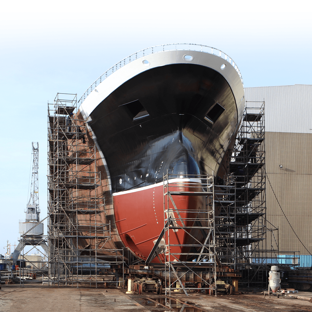 Front view of a large ship hull in dry dock, surrounded by metal scaffolding for construction or maintenance. The hull is painted black and red, with an industrial shipyard building and crane visible in the background.
