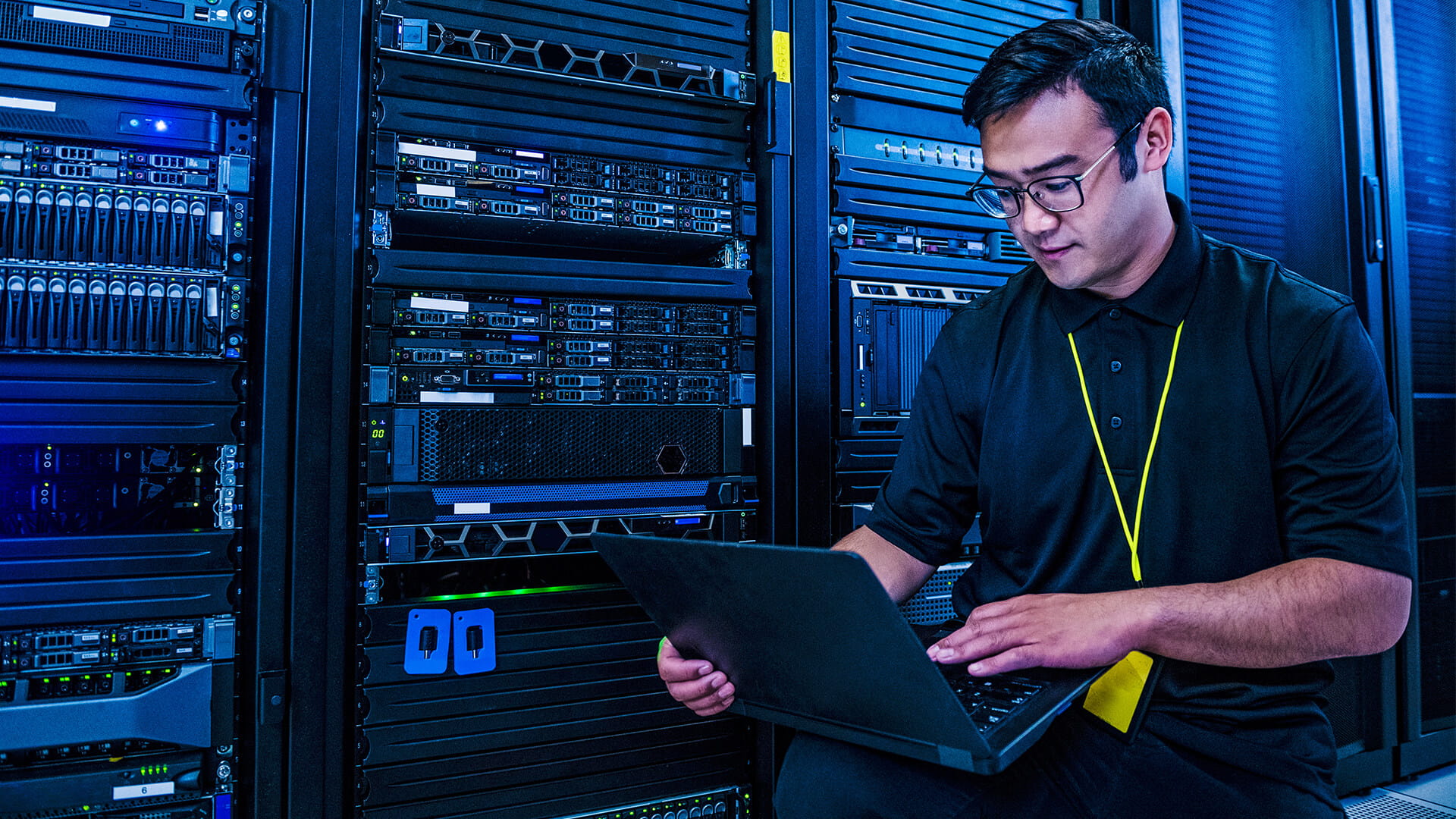 IT professional using a laptop while working in a server room with rows of data center racks.