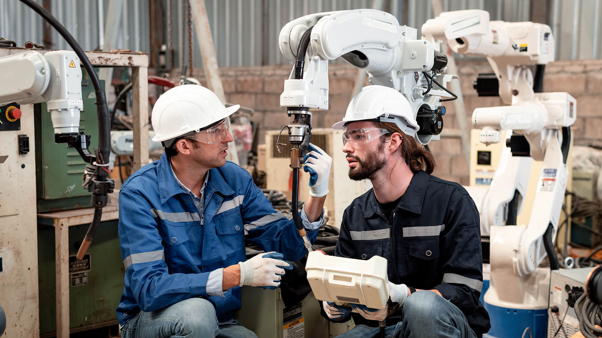 Two manufacturing workers wearing safety gear inspect and program an industrial robotic arm inside a factory.