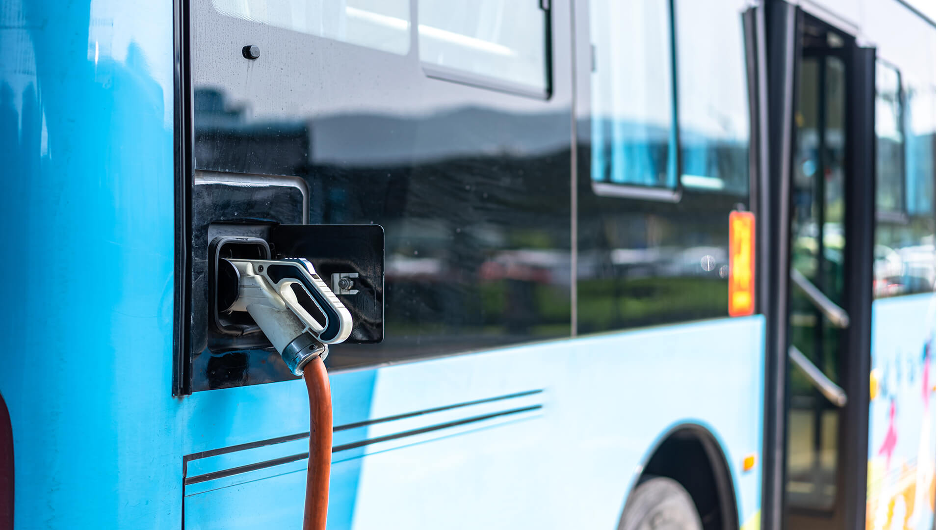 Close-up of an electric charging plug connected to the side port of a blue public transit bus, with the bus door and wheel visible in the background.