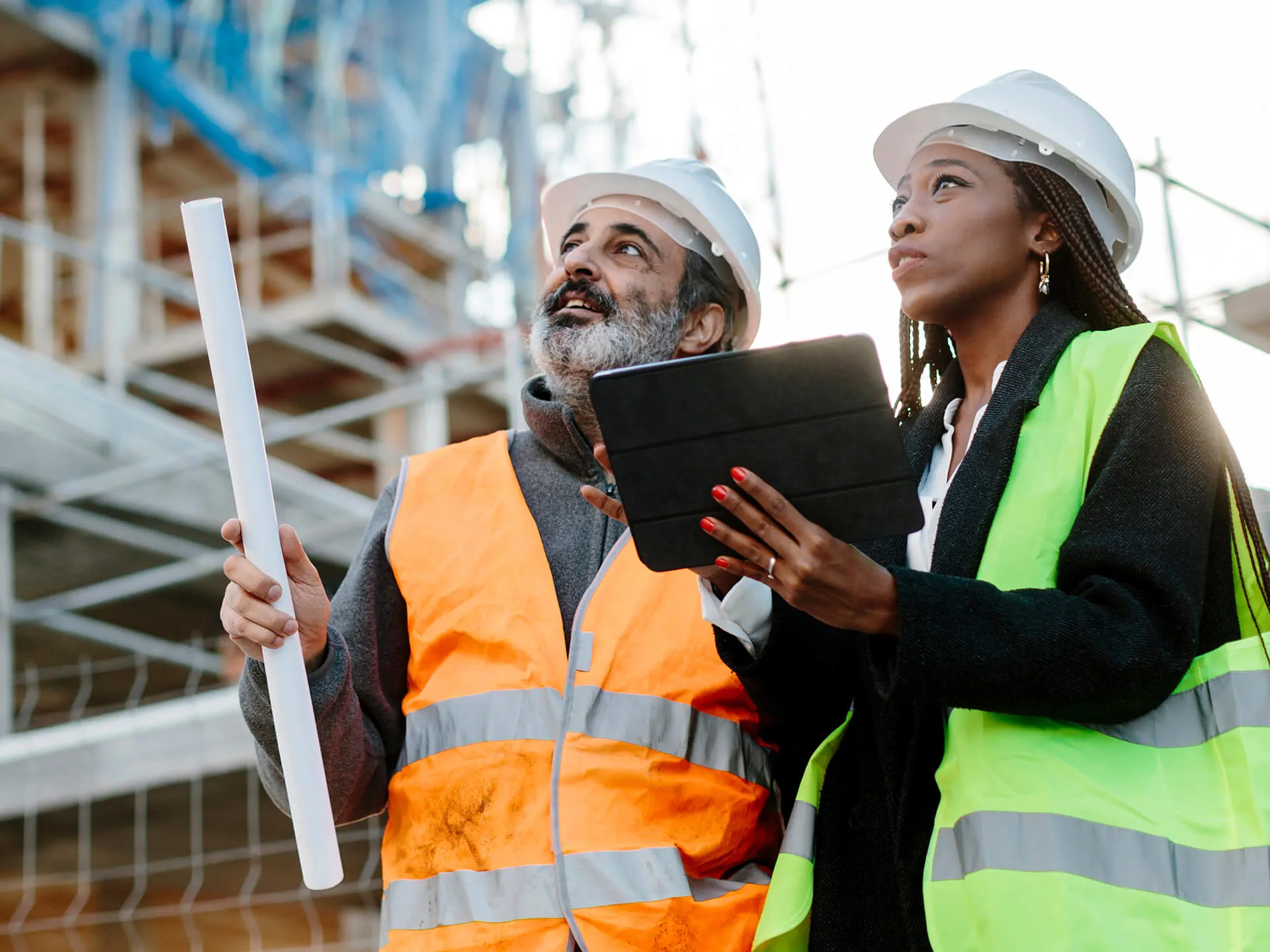 Two construction professionals wearing hard hats and high-visibility safety vests review plans at an active construction site, with one holding rolled blueprints and the other using a tablet while both look upward toward scaffolding.