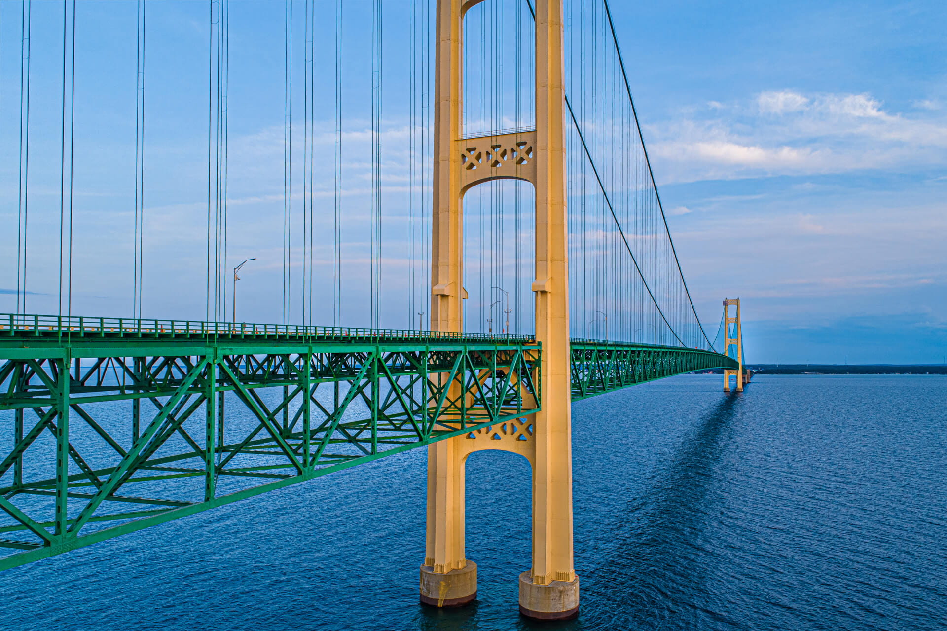 The Mackinac Bridge stretches across blue water, with green steel trusses and tall tan suspension towers receding into the distance under a clear sky.