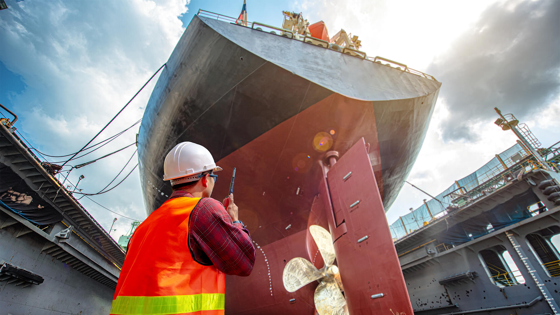 Worker in a high-visibility orange safety vest and white hard hat inspecting the underside and propeller of a large cargo ship in a dry dock under a cloudy sky.