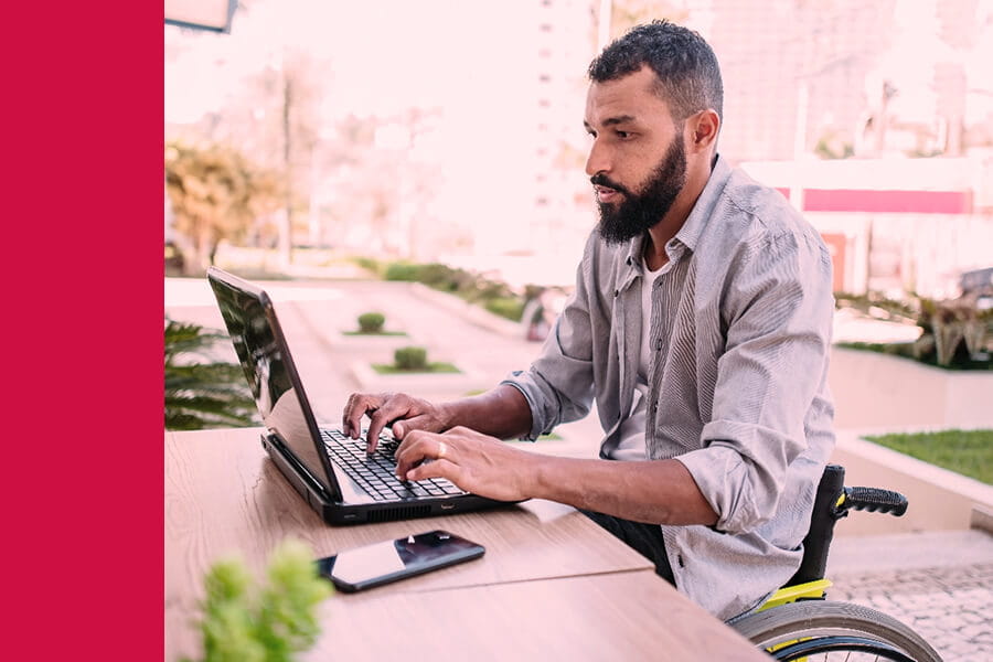 An adult man with a medium-light complexion seated in a wheelchair and working on a laptop outside on a bright sunny day.