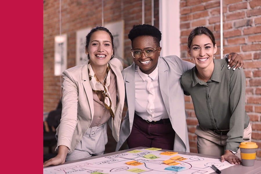 An ethnically diverse group of three coworkers smiling and embracing with arms around each other in an industrial-themed office space.