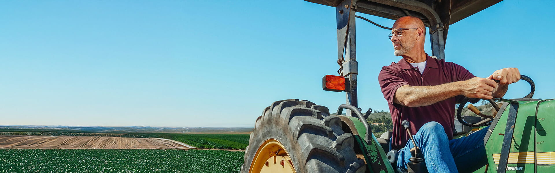 A farmer driving a green tractor in a vast agricultural field under a clear blue sky, looking to the side with a focused expression. The scene highlights rural farming and agricultural work in a serene outdoor environment.