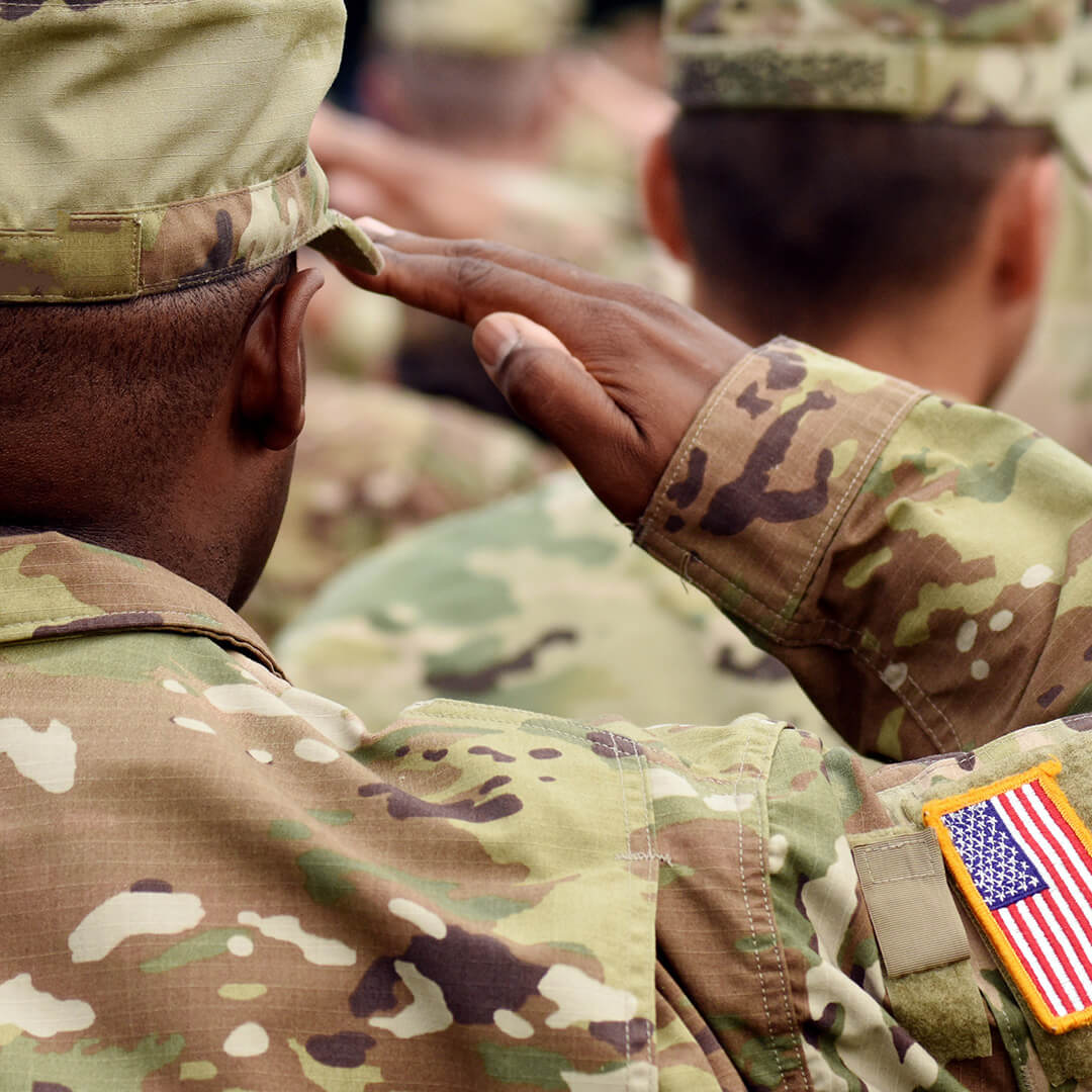 A Black military veteran dressed in a camouflage uniform with back turned and saluting in a line of other veterans.
