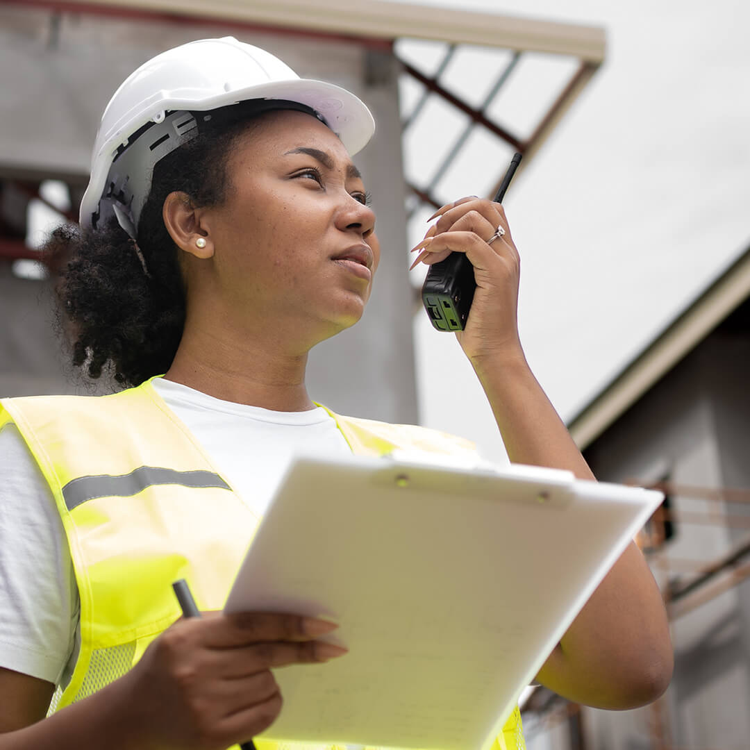 A dark-skinned woman dressed in a construction vest and hard hat holding a clipboard and talking into a handheld transceiver.