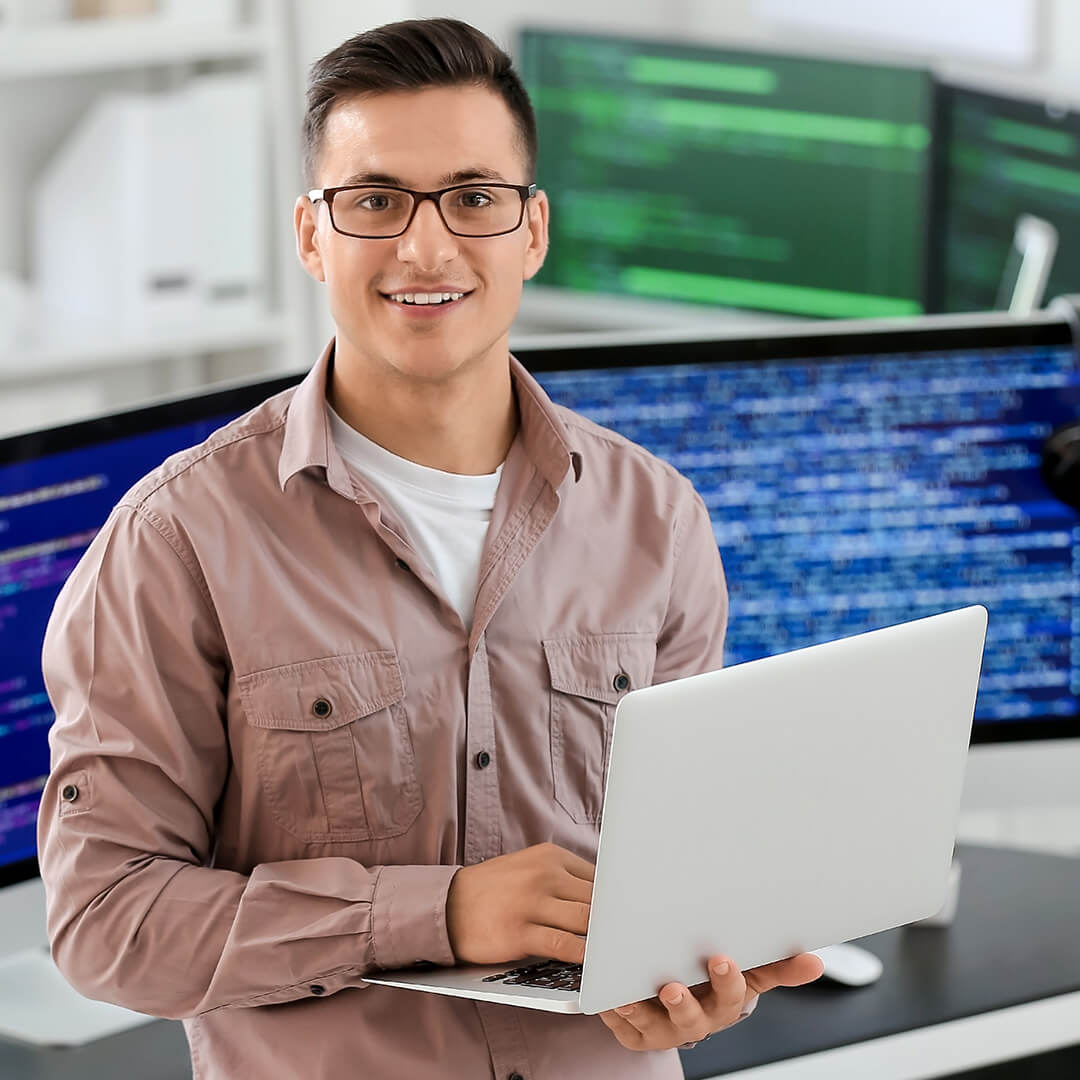 A young white computer programmer wearing glasses and smiling while holding a laptop with computer monitors displaying code in the background.