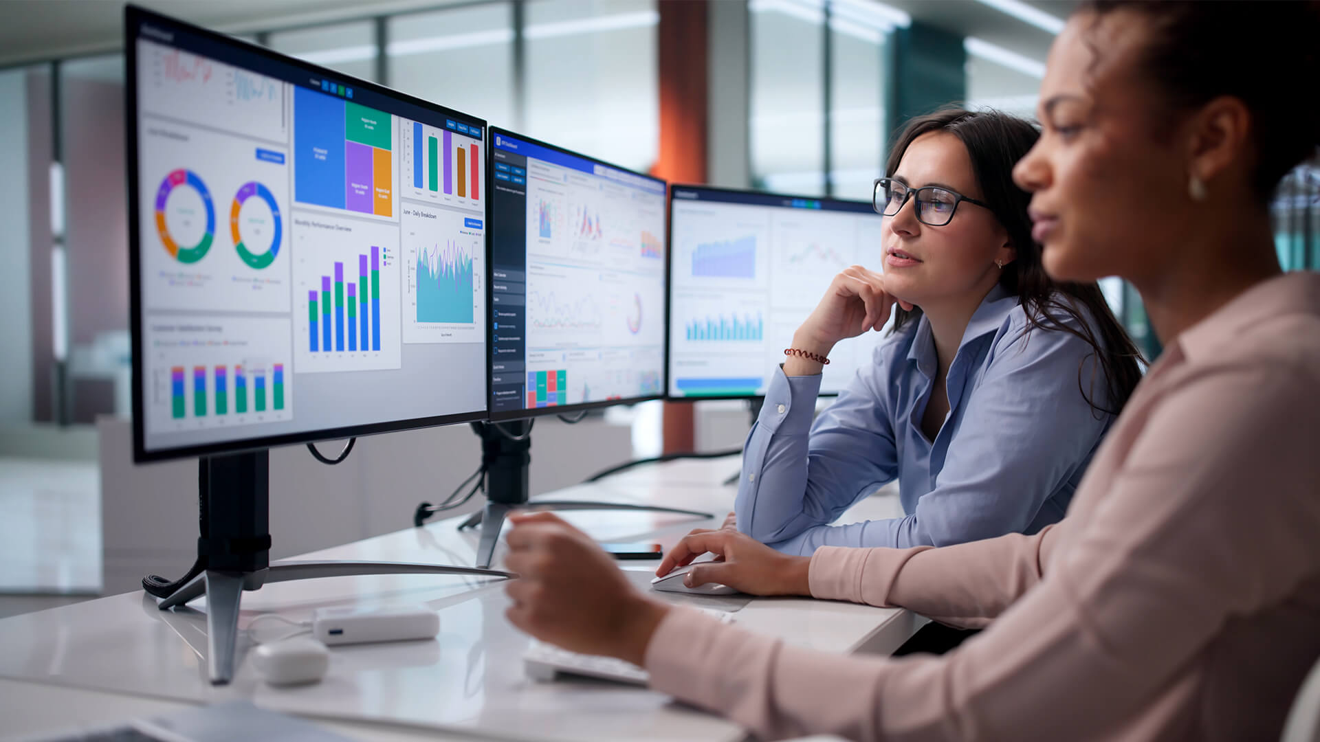 wo individuals sit at a desk reviewing charts and graphs on multiple computer monitors.