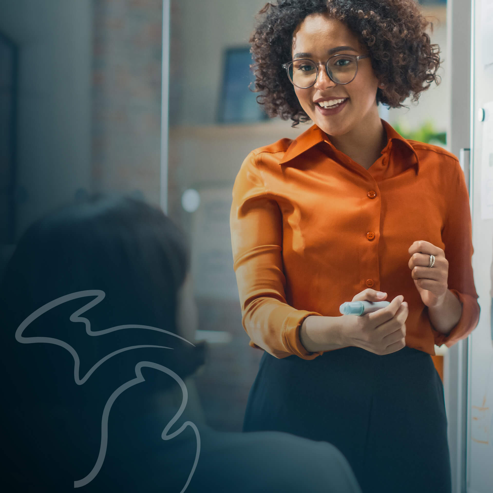 An company director smiling while standing at a dry erase board with a marker in hand and conducting a brainstorming session with members of her team.