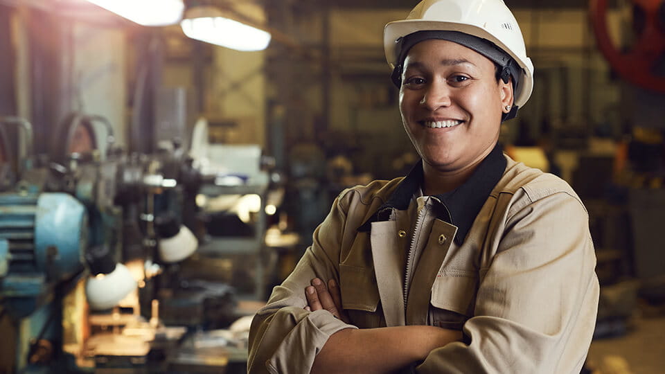 A shop apprentice wearing a safety hat and coveralls smiling with arms crossed.