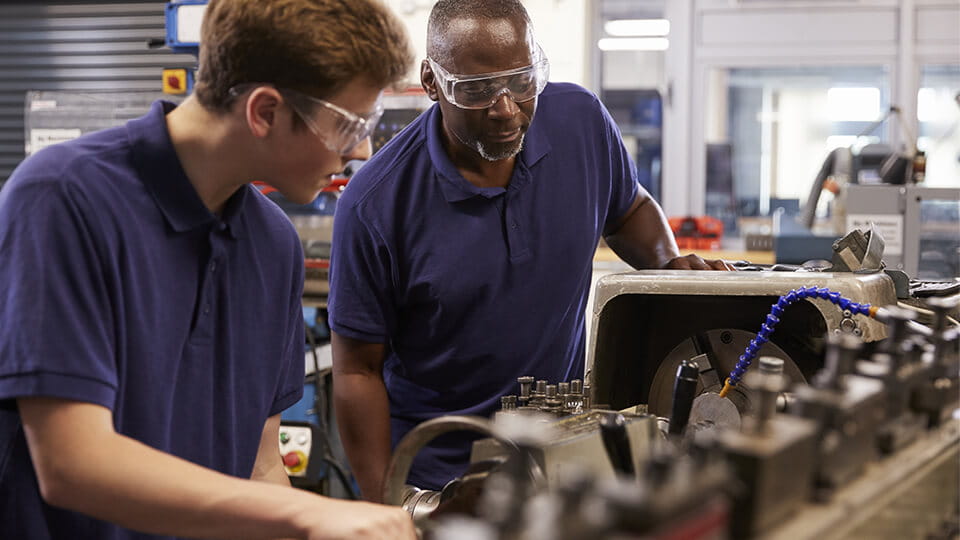 A manufacturing apprentice working with an experienced tradesperson to learn how to use a metal factory lathe.